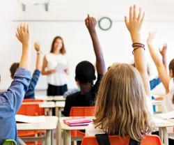 Imagem de alunos, sentados numa sala de aula, com o braço no ar para responderem à professora.